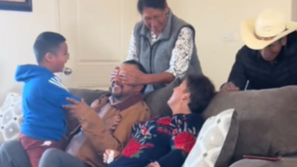 A man and woman sit on a couch. A kid sits on the man's lap. Standing behind the couch are a woman and a man. The woman covers the eyes of the man sitting on the couch. They both smile.