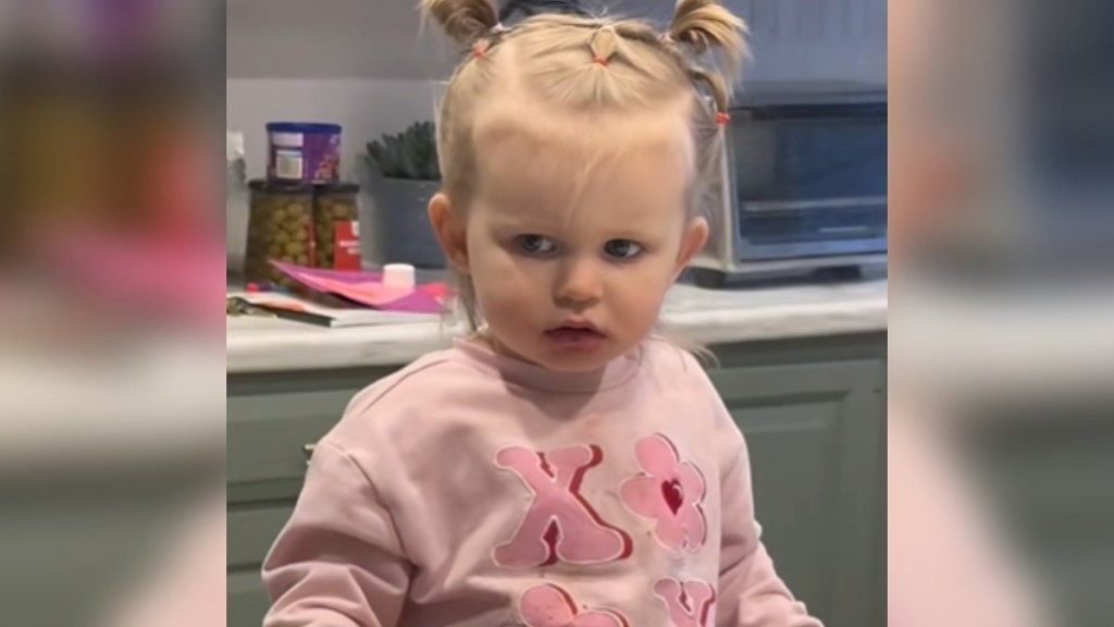 A toddler sits at a kitchen counter. She looks baffled while concentrating on something that can't be seen in this image