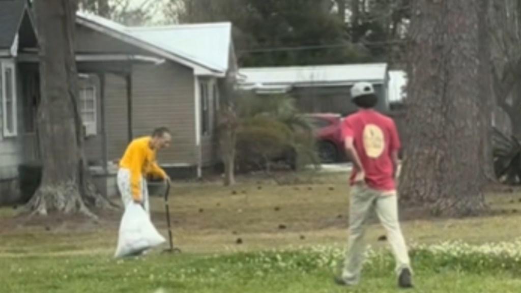 From a distance, a young man can be seen walking across a yard and toward an elderly woman. She walks with a cane in one hand and a trash bag in the other