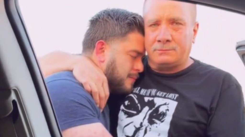 Close up of a man and his adult son embracing. Dad looks at the camera, tears in his eyes