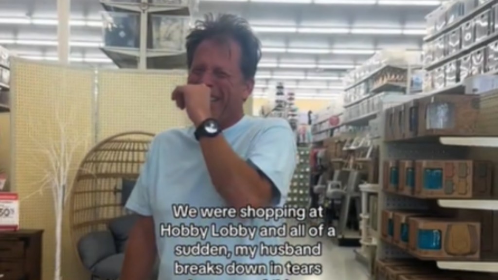 A man stands in a Hobby Lobby, laughing hysterically as he uses a hand to cover his mouth. Text reads: We were shopping at Hobby Lobby and all of a sudden, my husband breaks down in tears