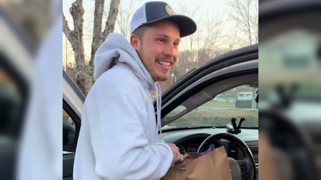 Close up of a man standing outside of a car, the door open. He smiles wide as he holds a brown bag in his hands.