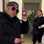 Two women stand in a hallway. One is close to the camera while the other is farther down the hall. The closer woman wears a hat backwards and sunglasses. She holds up a small flashlight and shines it down at someone we can't see. The woman in the back also wears a hat backwards. She has her arms crossed and holds a paper towel roll in one hand.