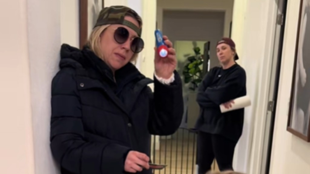 Two women stand in a hallway. One is close to the camera while the other is farther down the hall. The closer woman wears a hat backwards and sunglasses. She holds up a small flashlight and shines it down at someone we can't see. The woman in the back also wears a hat backwards. She has her arms crossed and holds a paper towel roll in one hand.