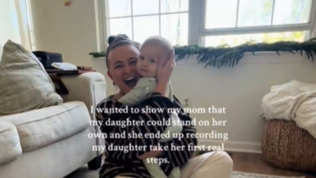 A woman sits on the floor of a living room. She holds her baby closer to her face, smiling wide with her mouth open. The baby looks a bit confused but is happy to be there. Text on the image reads: I wanted to show my mom that my daughter could stand on her own and she ended up recording my daughter take her first real steps