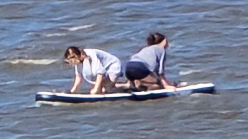View from a distance of two teen girls out on the water on paddleboards. They face away from each other and are resting on their knees and hands