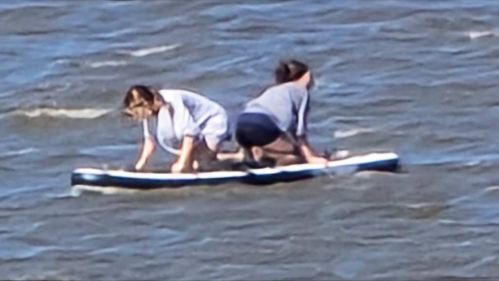 View from a distance of two teen girls out on the water on paddleboards. They face away from each other and are resting on their knees and hands