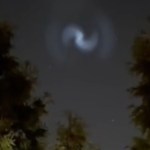 View of the night sky. Large trees can be seen all around. In the sky, a large spiral of light can be seen