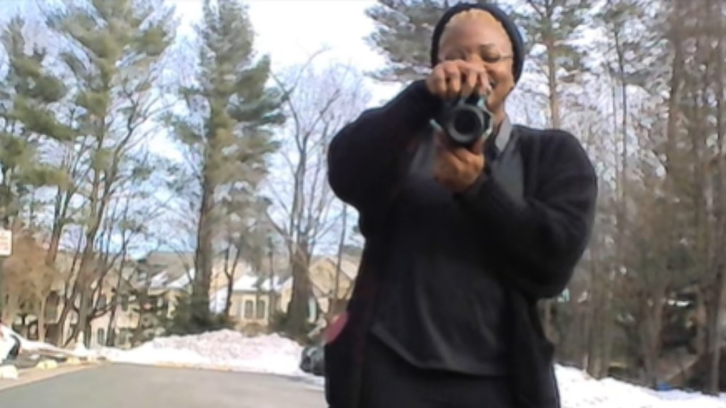 View of a woman standing outside, camera in hand. She's smiling wide as she aims the camera the person taking her photo. The perspective is from a toddler