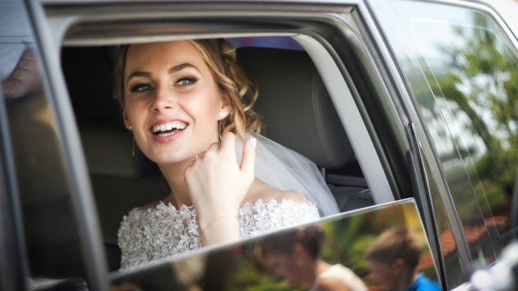 A bride sits in a car, window rolled down. She smiles, a look of surprise in her face. Her mouth is agape as she looks at something in the distance that can't be seen in this image
