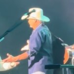 Alan Jackson stands on stage near a mic. He holds open his arms for his wife who smiles as she approaches