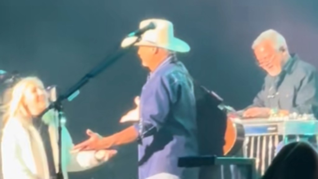 Alan Jackson stands on stage near a mic. He holds open his arms for his wife who smiles as she approaches