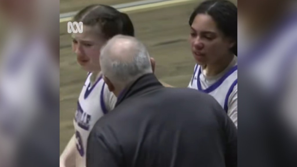 View of a man from behind as he approaches a teen basketball players. That teen is crying and looks afraid. A teammate stands next to her. This second teen looks angrily at the man and is starting to speak