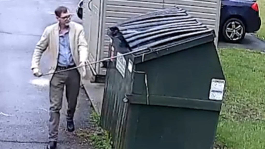 A man holds a stick, using it as he starts to open the dumpster next to him. Something dark is peaking out of the dumpster