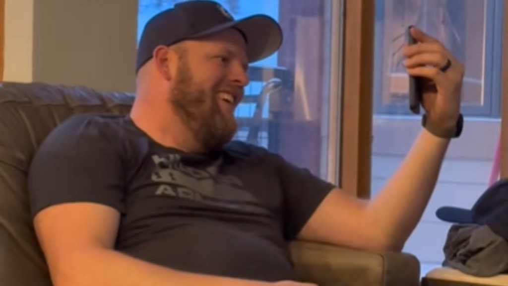 Close up of a man sitting on a couch. He holds up his phone, talking to someone on a video call. He smiles wide, mouth slightly open, as he looks at the phone