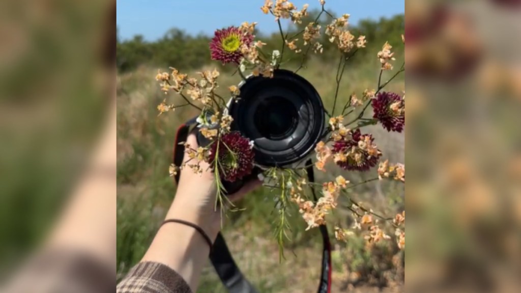 A woman holds out a professional looking camera while standing in a field. Flowers are attached to the camera and extend past the lens