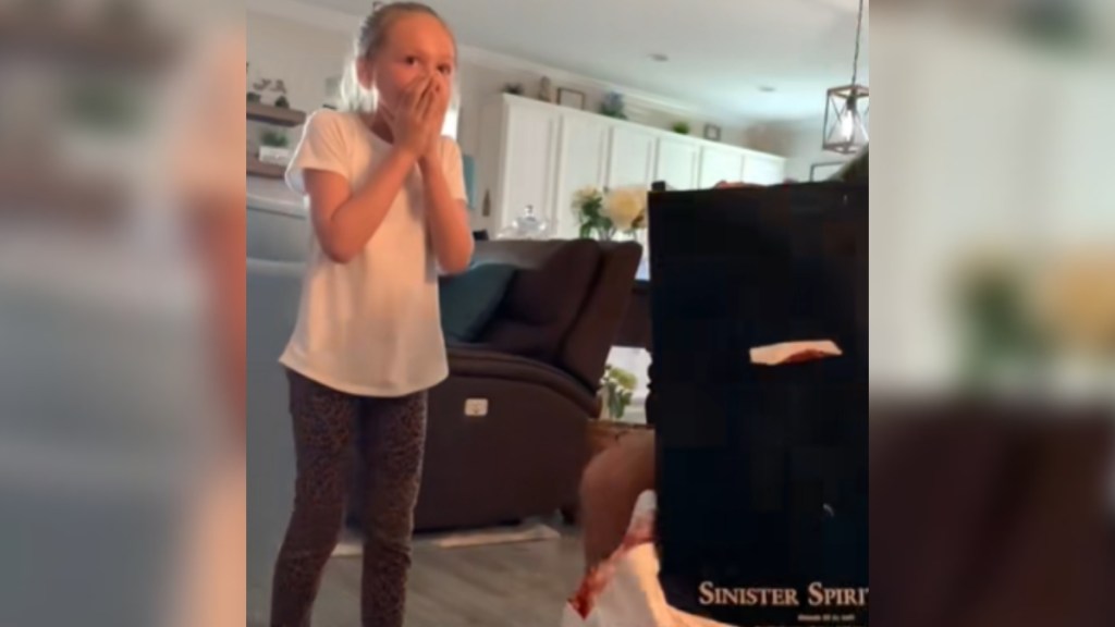 A little girl stands with a shocked expression on her face - her eyes are wide and she covers her mouth with her hands. She's standing near a box. The back of the box is toward the camera. On the back reads: Sinister Spirit