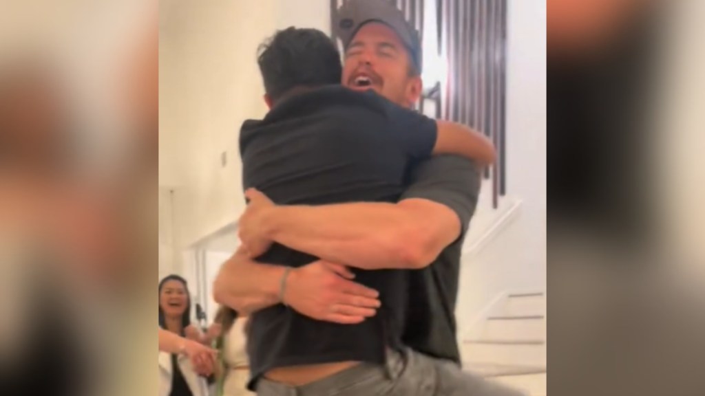 A large man smiles wide as he holds a smaller man in his arms. The shorter man has his legs wrapped around the taller man.