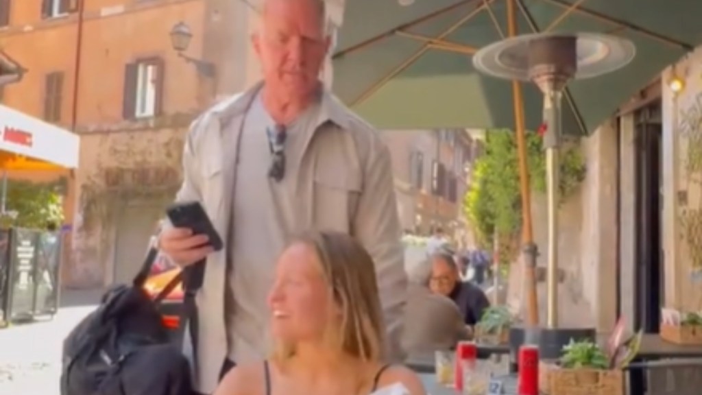 A woman sits at an outdoor dining table. She smiles as she starts to look to the side. A man stands behind her, slowly approaching