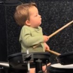 A 19-month-old boy sits at a drum set, sticks in hands, as he plays