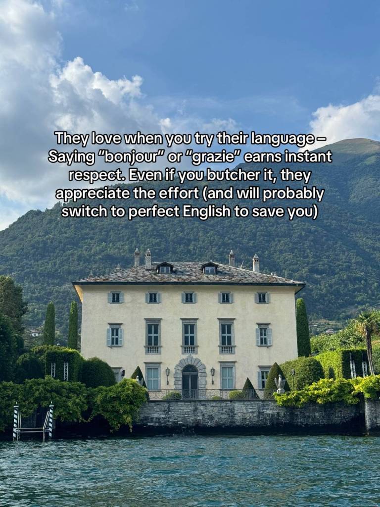 A giant home sits feet away from water. At a distance behind the home are giant mountains. Text on the image reads:

They love when you try their language - Saying "bonjour" or "grazie" earns instant respect. Even if you butcher it, they appreciate the effort (and will probably switch to perfect English to save you)