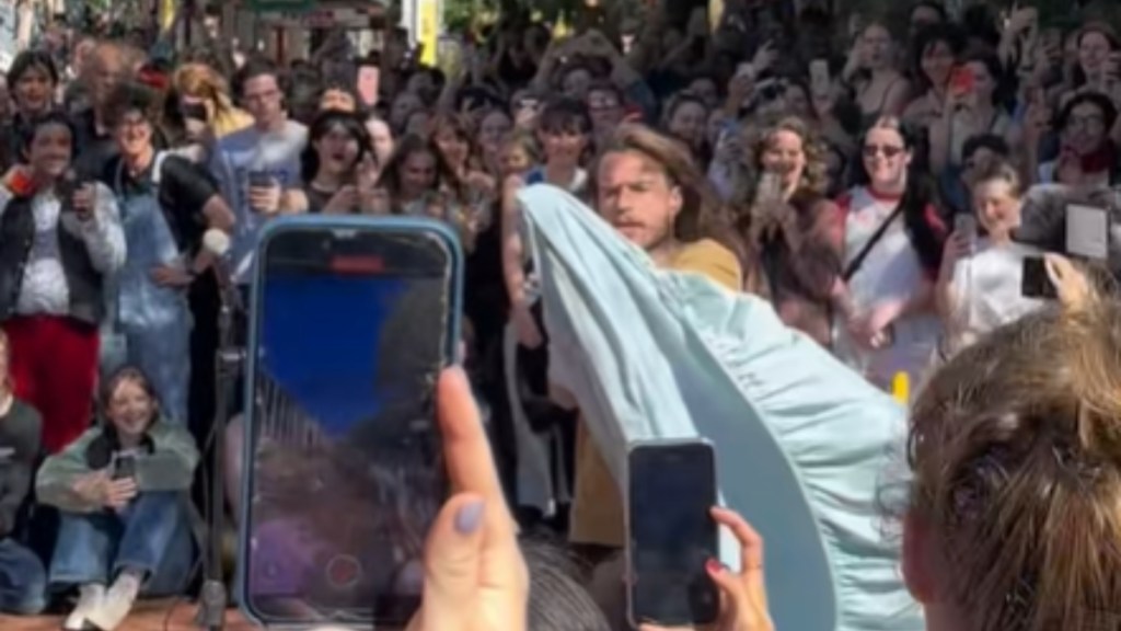 A massive crowd stands around one man. This man holds up a fitted sheet as he demonstrates how to fold it