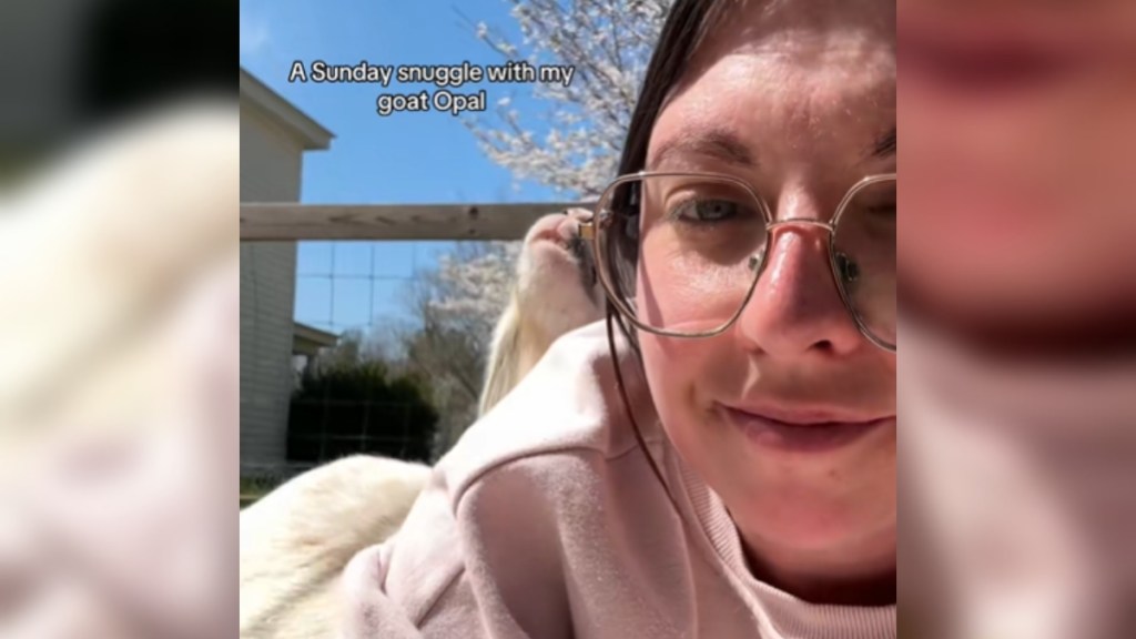 Close up of a woman as she lays on the ground outside. A small part of a goat's face can be seen peaking out from behind her shoulder. Text on image reads: A Sunday snuggle with my goat Opal