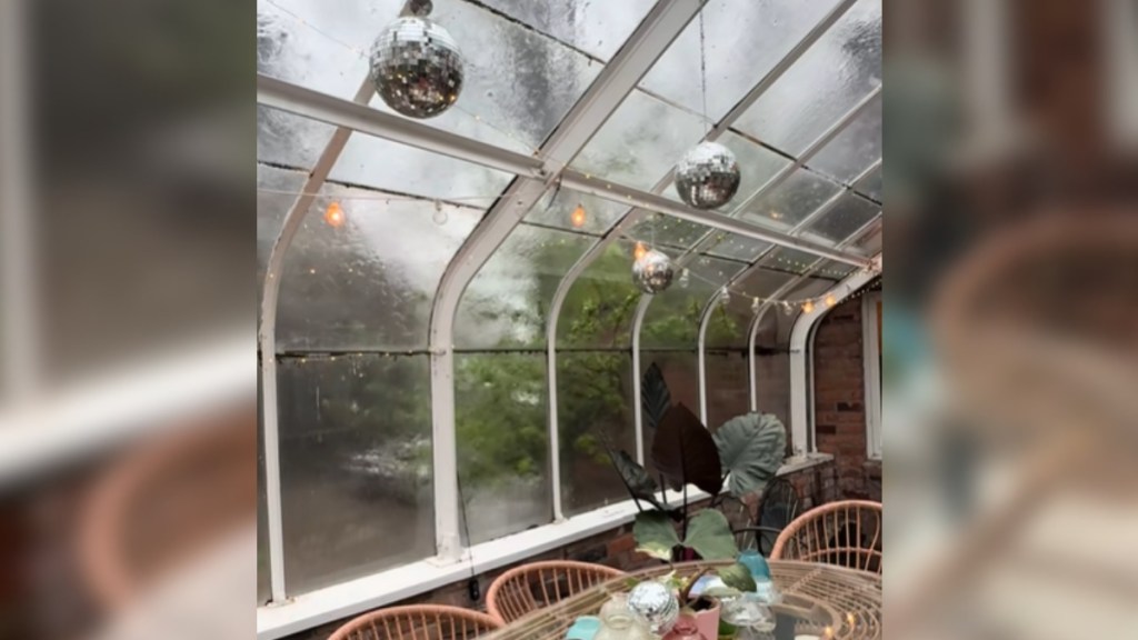 View from inside a greenhouse. There's an outdoor dining table with chairs. Fairy lights and two disco balls decorate the ceiling area. Rain gently falls on the glass wall and ceiling