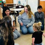 A group of high school students sits in a circle around a little boy, smiling and chatting