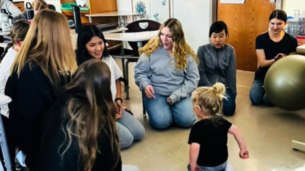 A group of high school students sits in a circle around a little boy, smiling and chatting