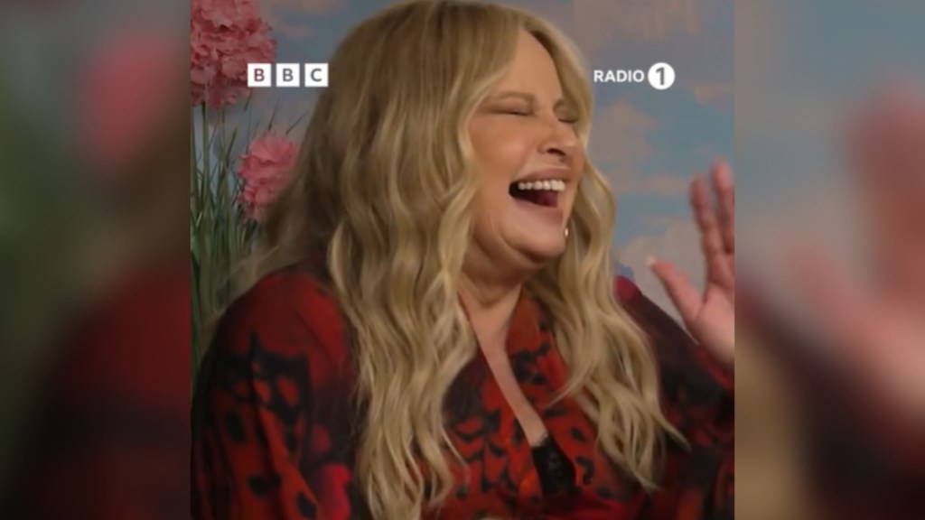 Close up of Jennifer Coolidge as she laughs so hard, her eyes are closed and her mouth is wide open. One hand is raised in the air.