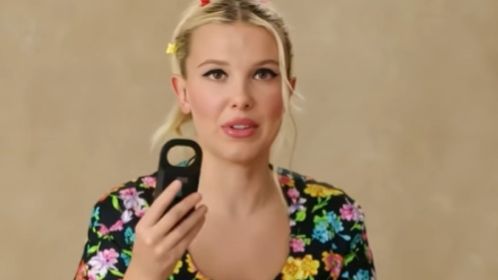 Close up of Millie Bobby Brown holding up a microchip scanner as she talks