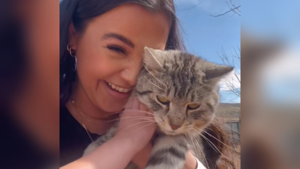 Close up of a woman as she holds a cat closely to her face. The woman smiles wide, emotional. A kid's hand can also be seen petting the cat.