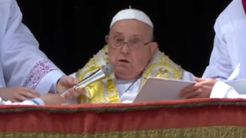 Pope Francis speaks into a mic being held for him. Another person holds a paper that he's reading from.