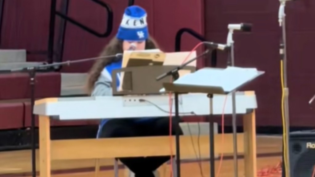 View from a distance of a teen girl sitting behind a piano. She wears a hat, and her face is partially covered by a mic and a music booklet sat in front of her