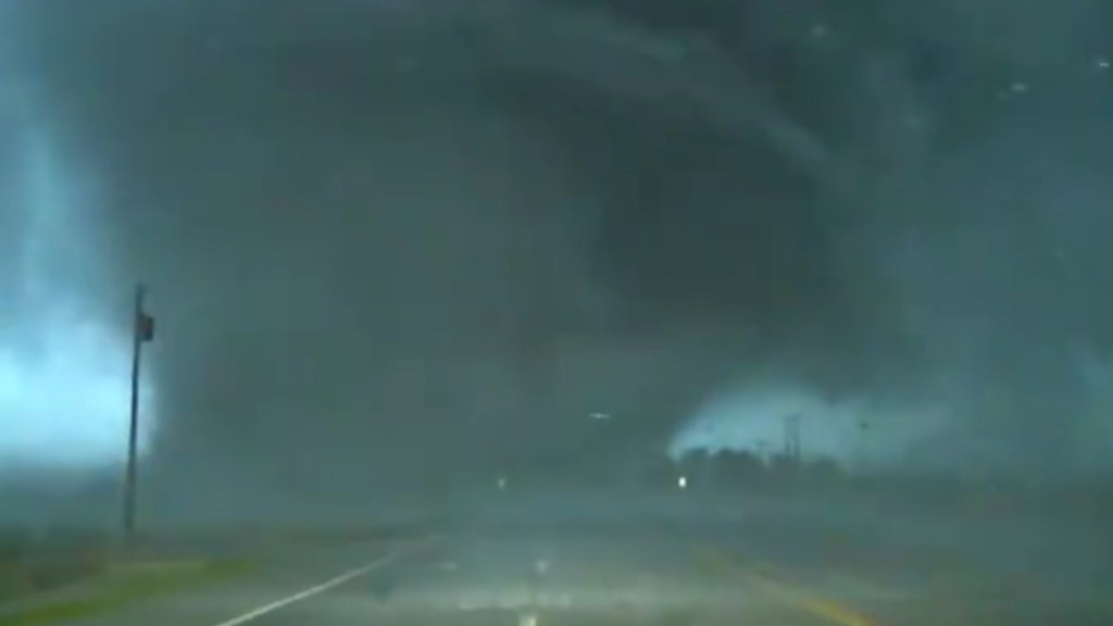 In the distance, a massive tornado makes its way across the road, debris flying everywhere