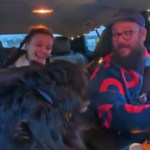 View of the interior of a car. The man in the driver's seat smiles as he turns to look in the back seat at the woman there. A big black dog sits in the passenger seat, looking back at the woman who is smiling at him.
