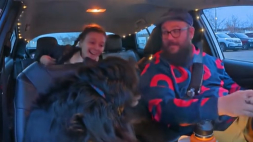 View of the interior of a car. The man in the driver's seat smiles as he turns to look in the back seat at the woman there. A big black dog sits in the passenger seat, looking back at the woman who is smiling at him.