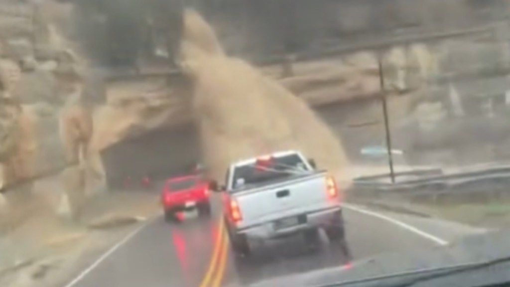 View from inside a vehicle. Two other vehicles are in front of them. A massive, murky waterfall cascades down on the road in the distance
