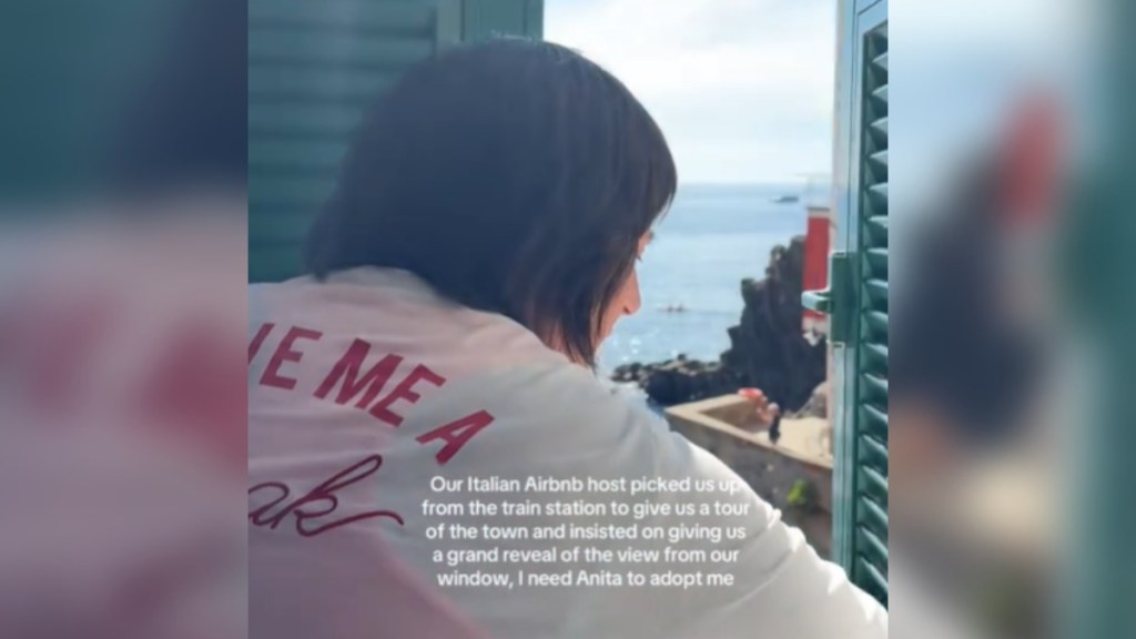 View from behind of a woman as she opens a window, partially revealing the stunning view. Text on the image reads: Our Italian Airbnb host picked us up from the train station to give us a grand reveal of the view from our window, I need Anita to adopt me