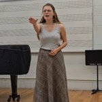 A young woman stands near a piano in a classroom. As she sings, she stretches out an arm in front of her as if reaching. She looks emotional