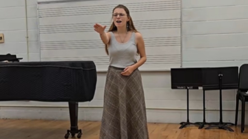 A young woman stands near a piano in a classroom. As she sings, she stretches out an arm in front of her as if reaching. She looks emotional