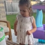 A little girl stands in her backyard. She looks down at the butterfly that's starting to fly toward her
