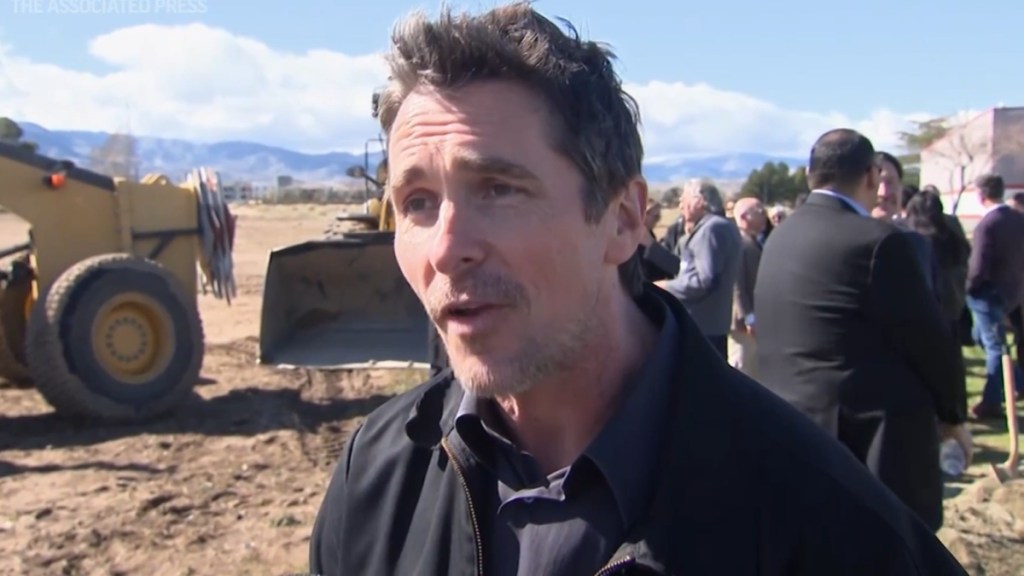 Christian Bale stands outside, talking to the camera. Construction is happening nearby behind him. A group of people also stand at a distance