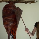 A young woman stands next to a hanging art piece that resembles a butterfly chrysalis. The woman gives a sly smile as she uses a device to start and pull back part of the chrysalis