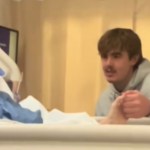 A woman lays in a hospital bed in a coma. A man sits next to her bed, holding her hand. He's singing