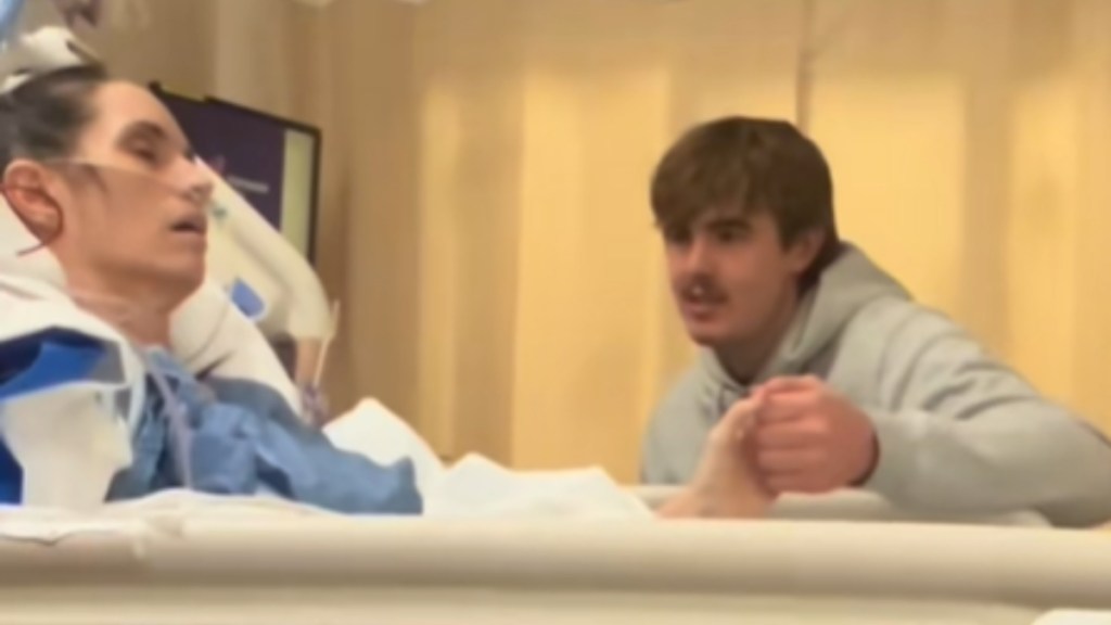 A woman lays in a hospital bed in a coma. A man sits next to her bed, holding her hand. He's singing