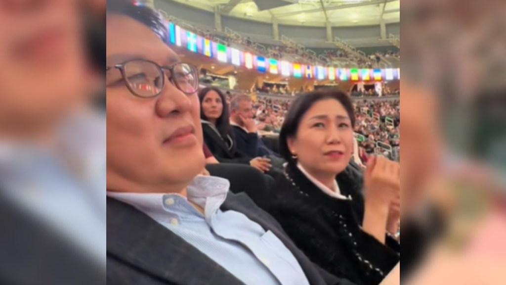 A dad and mom sit in a crowd at graduation day. Mom looks emotional as she looks over at the person capturing this moment. Dad stares forward, concentrating