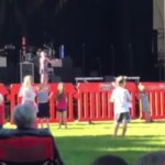 A small crowd gathers near a stage. On the stage, a little girl sings into a mic, one arm stretched out.