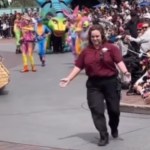 A Disney park parade is paused. The crowd looks on at a Disney employee who is walking up to something that can't be seen in this image, arms open wide
